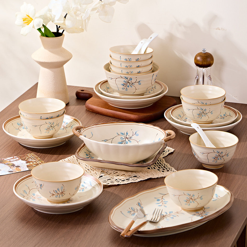 The set on a wooden table beside a flower vase: stacked rice bowls with spoons, a two-handled serving bowl, cups on saucers and an oval plate with a fork.