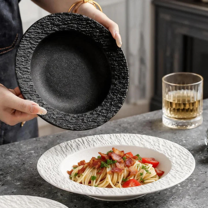 A person holds up a black textured-rim pasta bowl over a stone counter, with a white textured pasta plate of spaghetti in front.