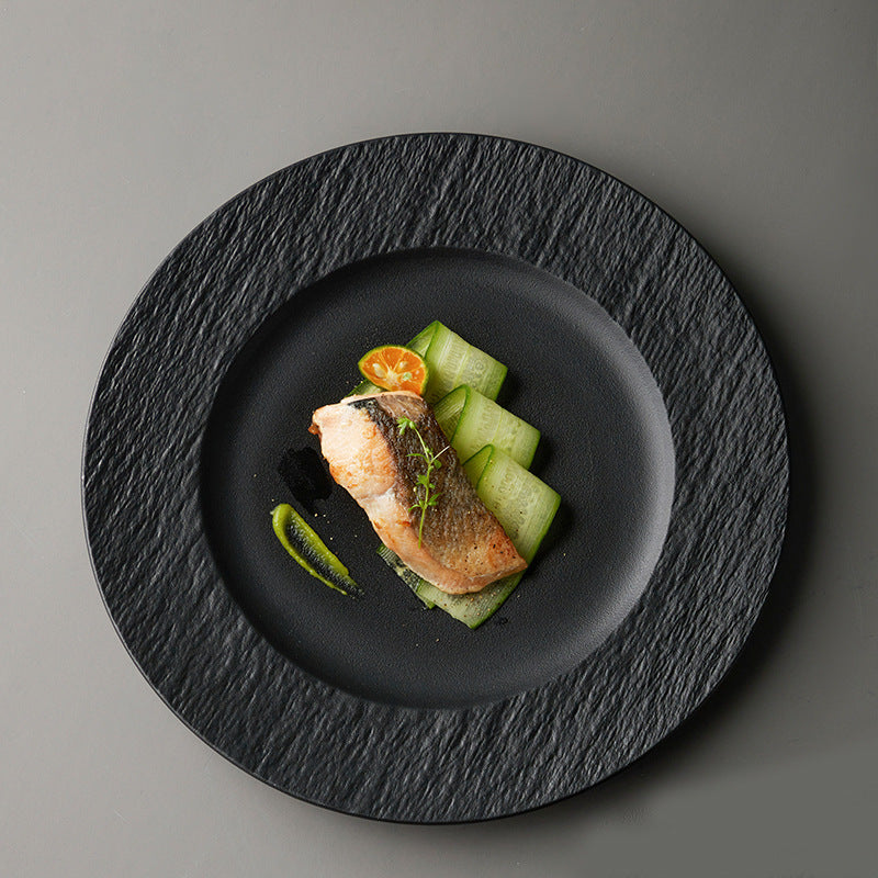 A black plate with the same textured rim seen from above, plated with a fillet of salmon and sliced cucumber.