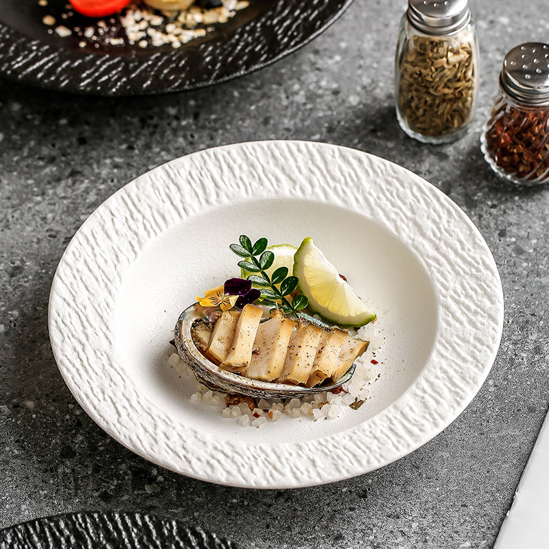A white textured-rim pasta plate holding grilled fish with a wedge of lime, on a grey stone counter beside spice jars.