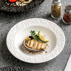 A white textured-rim pasta plate holding grilled fish with a wedge of lime, on a grey stone counter beside spice jars.
