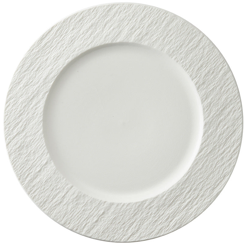 A wide white dinner plate on a white background, smooth centre well and a broad rim with a rough stone-like texture.