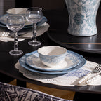 A place setting on a dark round table, a fluted bowl on layered blue-bordered plates over an embossed white charger, with two stemmed glasses and a floral vase behind.