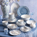 The full set displayed on a blue draped table beside a floral vase: stacked bowls, plates, an oval platter, a solid blue plate and a gravy boat.