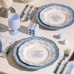 Two place settings on a cream cloth, each a floral-bordered plate over a solid blue plate, with a pale blue goblet and pearl-handled cutlery.