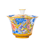 A lidded tea bowl on a white background, yellow ground covered in dense painted flowers in blue, pink and purple, with a gold rim and a gold band at the foot.