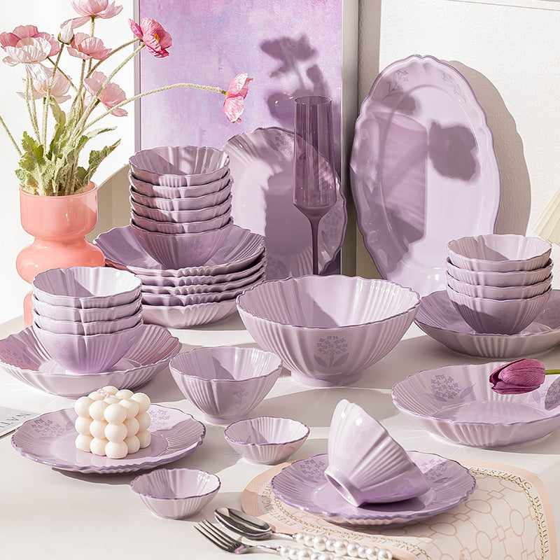 The lilac set laid out on a pale table beside pink poppies, with stacked plates and bowls, a large fluted serving bowl, small dishes and a spoon.