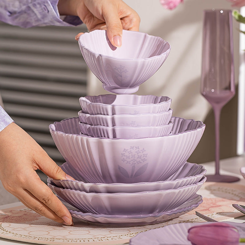 Two hands stack lilac bowls and plates on a table, the largest plate at the bottom and a small footed bowl on top.