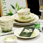 Stacked bowls and layered plates on a table with a printed card and a green glass, a leafy framed picture behind.