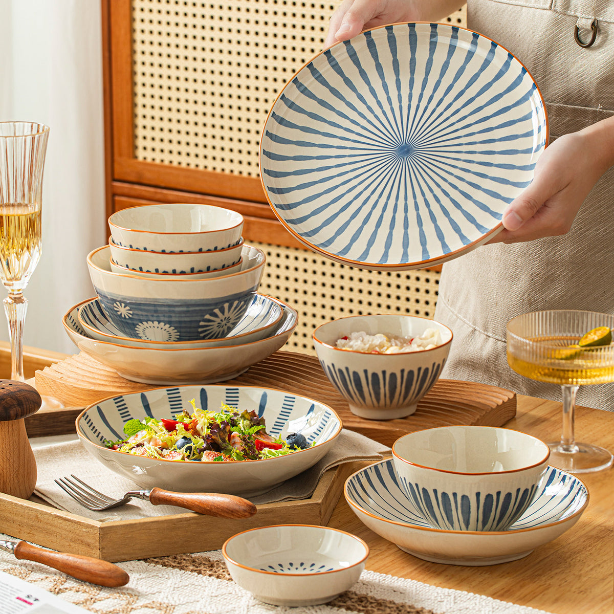 A person holds up a plate painted with radiating blue stripes above a wooden table set with matching bowls and a salad bowl.