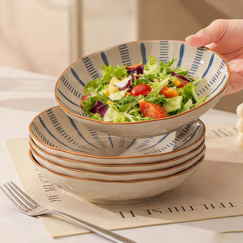 A hand steadies a striped salad bowl resting on a stack of matching bowls and plates on a cream mat.