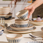 A hand lifts a small striped bowl from a stack of plates and bowls on a laid table.