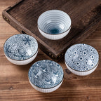 Four rice bowls in different blue and white patterns, fine stripes, crackle, small flowers and a wave scroll, on a wooden table beside a dark wooden tray.