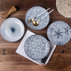 Four plates in different blue and white patterns laid on a wooden table with a magazine, a pepper grinder, gold cutlery and white chopsticks.