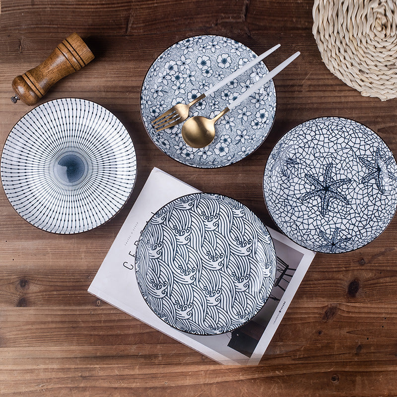 Four plates in different blue and white patterns laid on a wooden table with a magazine, a pepper grinder, gold cutlery and white chopsticks.