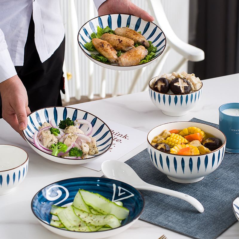 A person sets a marble table with the set: a wide leaf-pattern bowl of chicken wings, a small bowl of mushrooms, a soup bowl of corn and vegetables, a navy plate of sliced cucumber and a white soup spoon.
