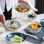 A person sets a marble table with the set: a wide leaf-pattern bowl of chicken wings, a small bowl of mushrooms, a soup bowl of corn and vegetables, a navy plate of sliced cucumber and a white soup spoon.