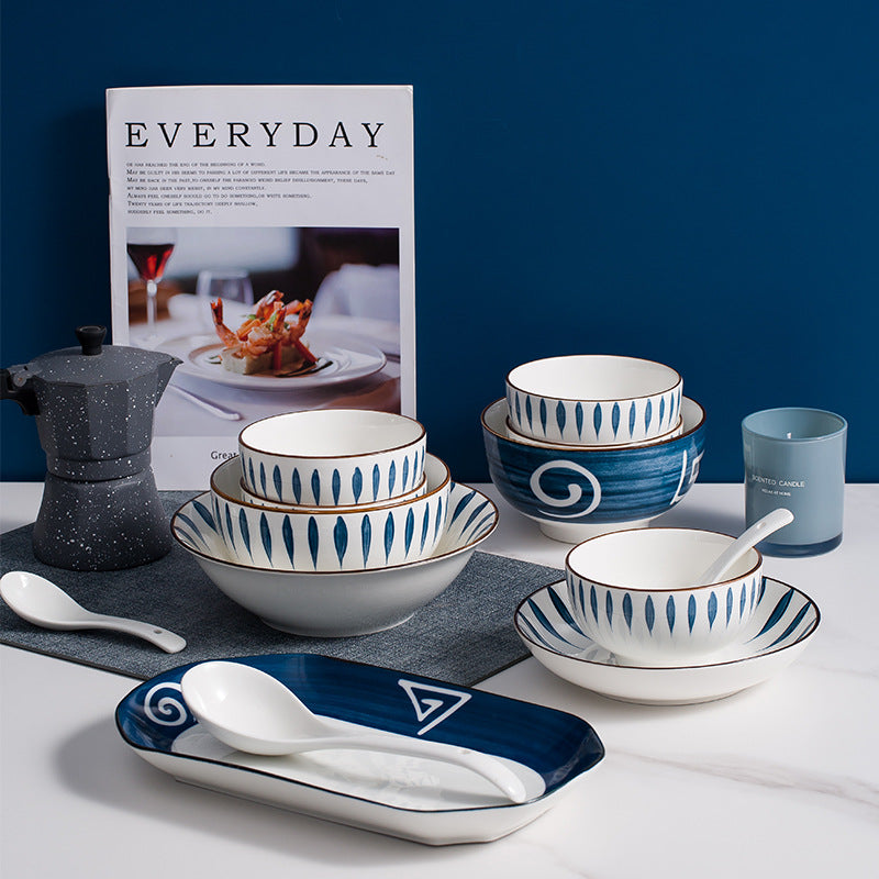 The pieces grouped against a deep blue wall: three leaf-pattern bowls in graduated sizes, a navy rice bowl with a painted spiral, a small saucer, and two white soup spoons resting on the oval navy serving dish.
