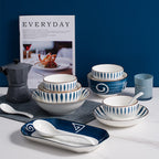 The pieces grouped against a deep blue wall: three leaf-pattern bowls in graduated sizes, a navy rice bowl with a painted spiral, a small saucer, and two white soup spoons resting on the oval navy serving dish.
