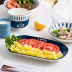 The oval navy serving dish holding diced pineapple, sliced tomato and lettuce, with a leaf-pattern rice bowl and a wide bowl behind it on a marble table.