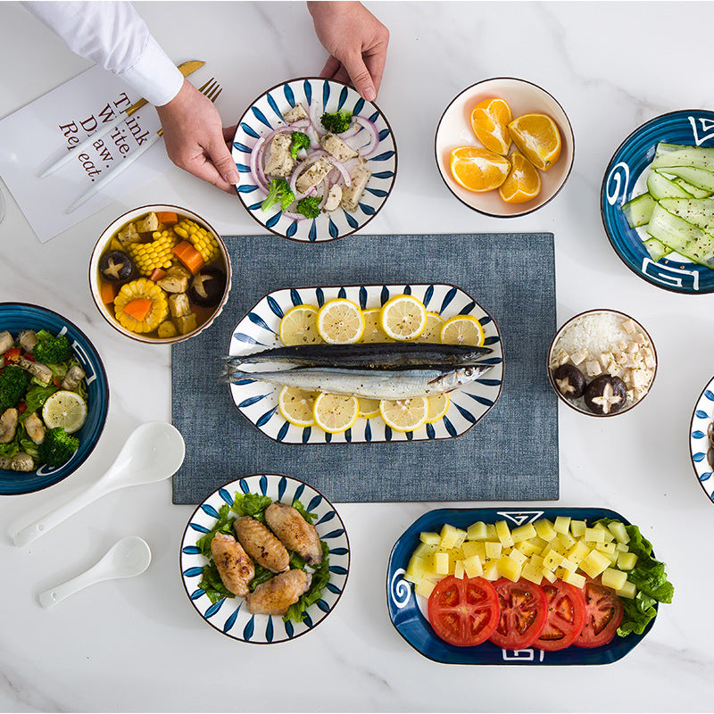 Overhead view of a laid table: leaf-pattern plates and bowls, the oval navy dish with grilled fish and lemon slices, small bowls of orange segments, rice and cucumber, and two white soup spoons.