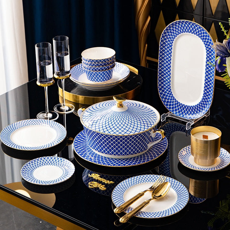 The set staged on a black and gold table: a lidded tureen, stacked bowls, an oval platter on a stand, plates and gold cutlery, with two champagne flutes.