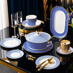 The set staged on a black and gold table: a lidded tureen, stacked bowls, an oval platter on a stand, plates and gold cutlery, with two champagne flutes.