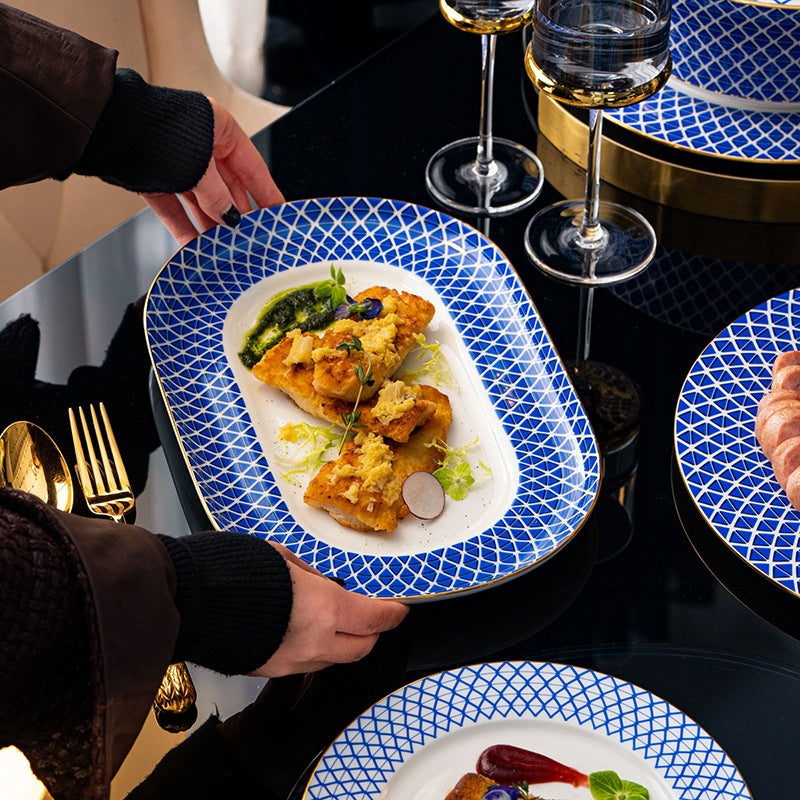 A person carries a rectangular platter of plated fish to a dark glass table set with the range.