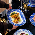 A person carries a rectangular platter of plated fish to a dark glass table set with the range.