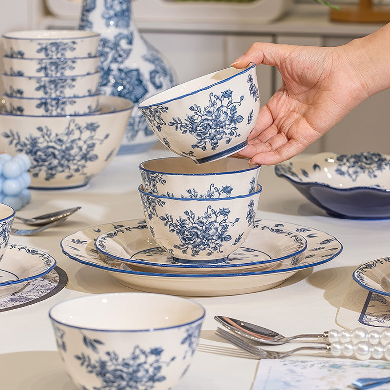 A hand lifts a floral bowl from a stack on a laid table, with more bowls, plates and a tall vase behind.