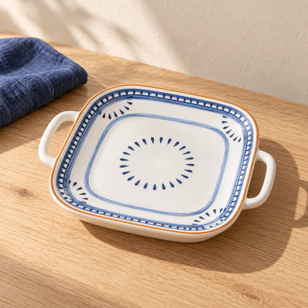 A blue rhyme square plate with two ear handles and a hand-painted blue motif, on an oak table with indigo linen.