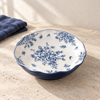 A blue and white 8 inch deep plate with a dense peony print and a deep indigo rim, on a pale limestone table in morning light.
