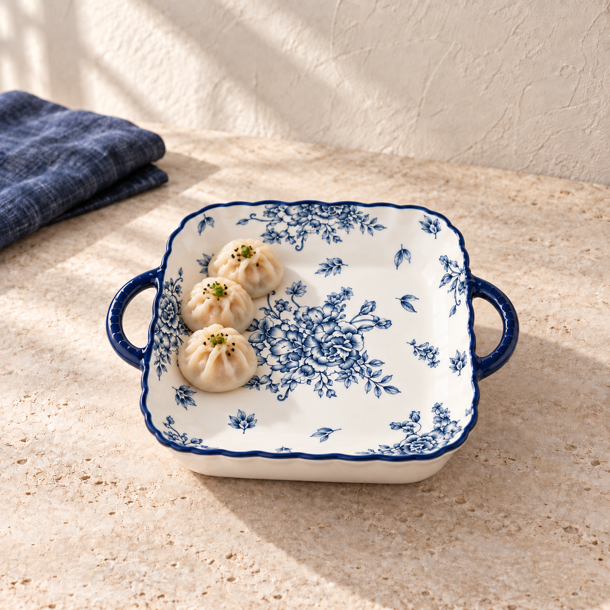 The blue and white eared square plate holding three steamed buns, on a pale limestone table. The food is styling and is not included.