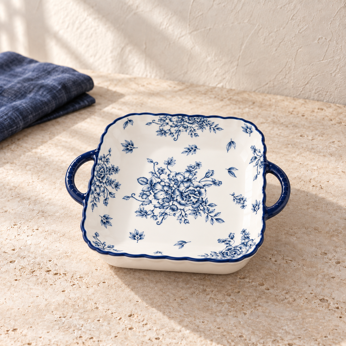 A blue and white square plate with two deep indigo ear handles and a peony print, on a pale limestone table in morning light.