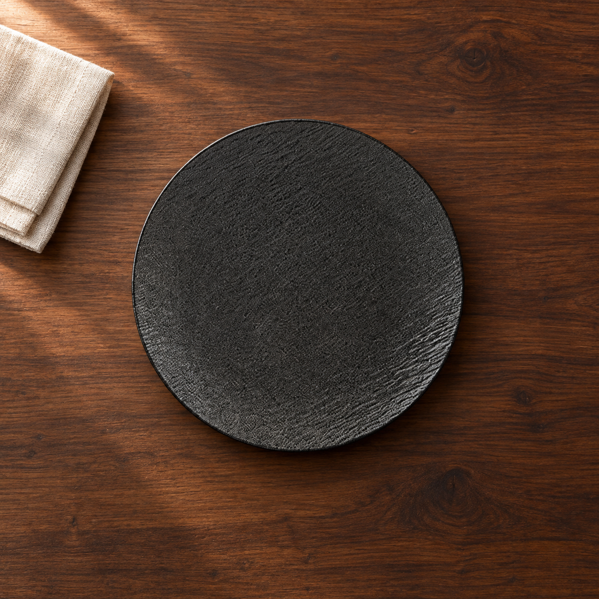 A matte black 10 inch shallow plate with a fine stone-like grain, on a dark walnut table beside a folded linen napkin, seen from above.