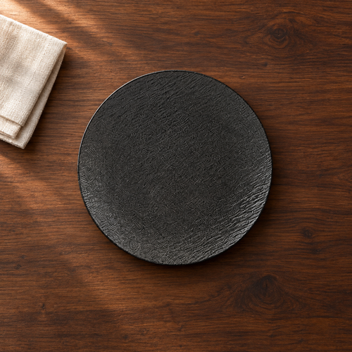 A matte black 10 inch shallow plate with a fine stone-like grain, on a dark walnut table beside a folded linen napkin, seen from above.