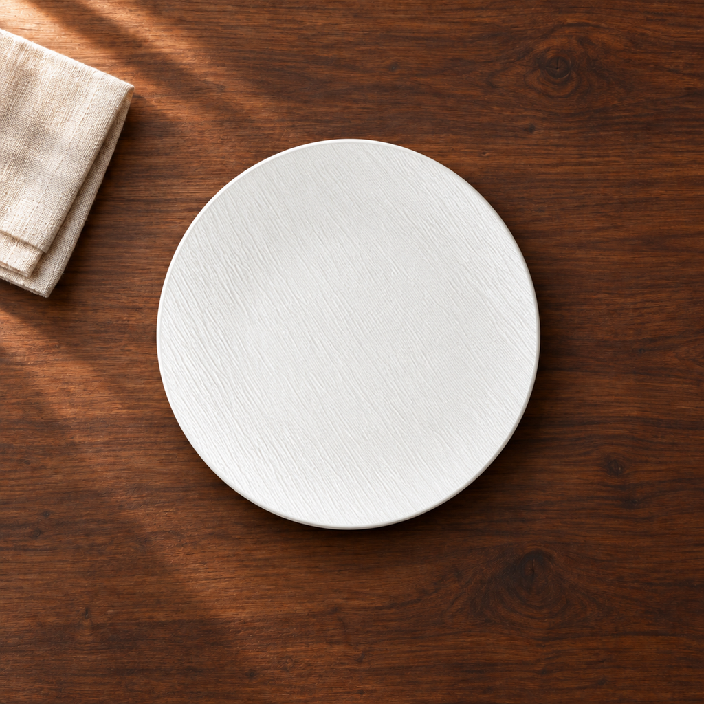 A matte white 10 inch shallow plate with a fine stone-like grain, on a dark walnut table beside a folded linen napkin, seen from above.