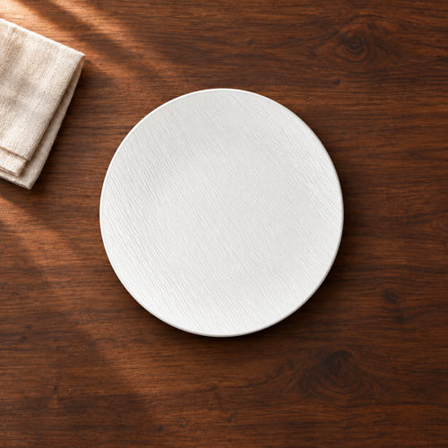 A matte white 10 inch shallow plate with a fine stone-like grain, on a dark walnut table beside a folded linen napkin, seen from above.