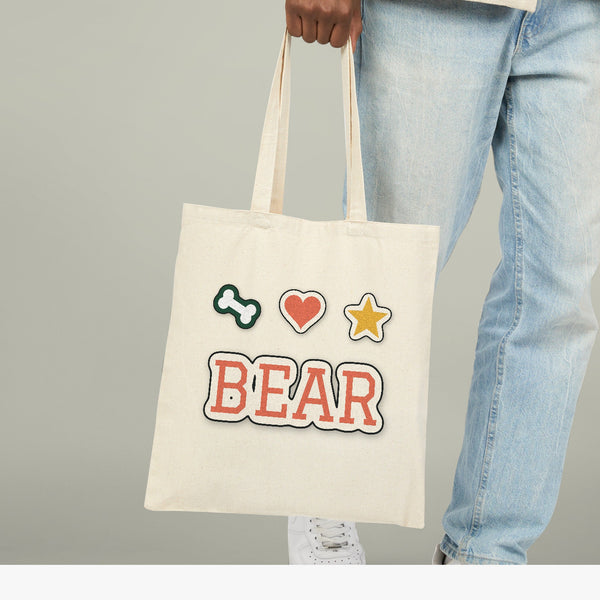 Natural cotton canvas tote carried by the handles, printed with the sample name BEAR in varsity lettering under a bone, a heart and a star
