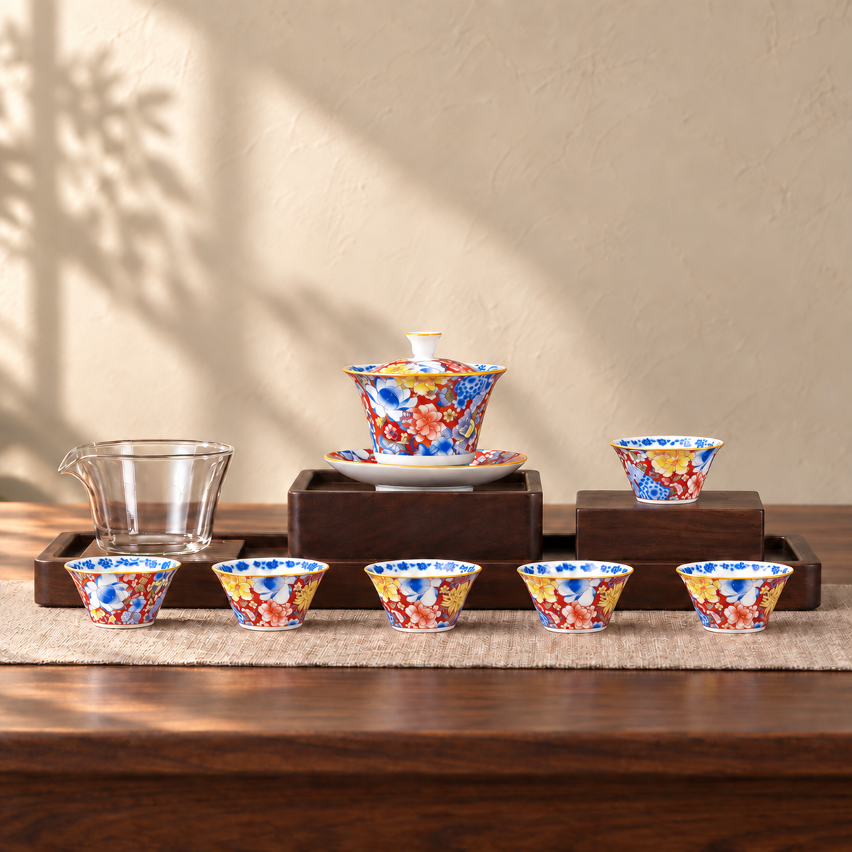 A tea set painted edge to edge in flowers on a red ground with gold rims: a lidded tea bowl on its saucer, six small cups and a clear glass pitcher. The wooden stand and mat are styling and are not included.