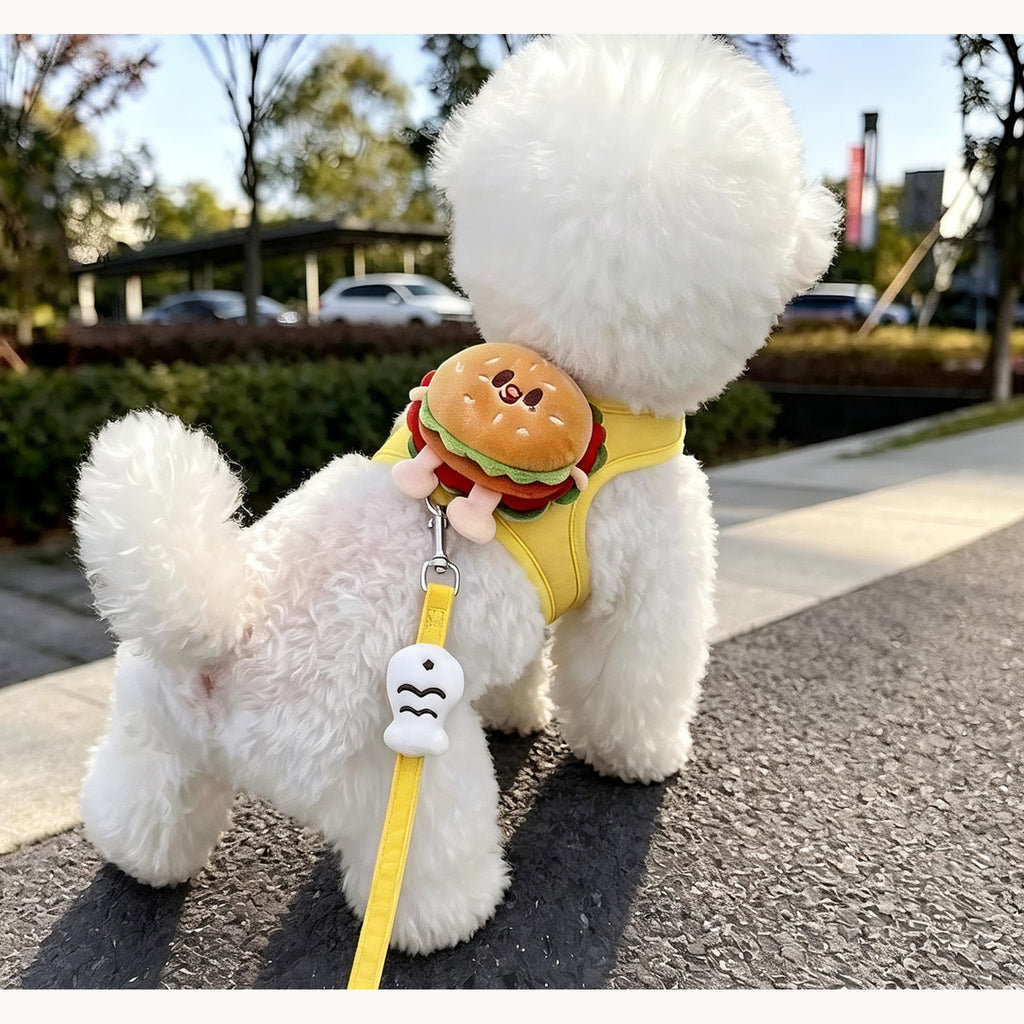 Small white dog walking away on a pavement in the yellow burger-motif harness and lead