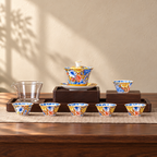 A tea set painted edge to edge in flowers on a yellow ground with gold rims: a lidded tea bowl on its saucer, six small cups and a clear glass pitcher. The wooden stand and mat are styling and are not included.